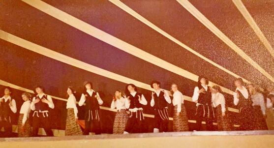 A group of dancers performing onstage, with the women wearing long dresses and headscarfs, and the men wearing black vests, hats, and trousers, white shirts, and tallit katan