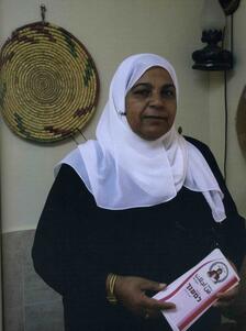 Amna Kenanalk holding a program, with a woven decoration hanging on the wall behind her