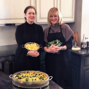 Two women standing in a kitchen, one holding a strainer with noodles and the other a strainer with greens