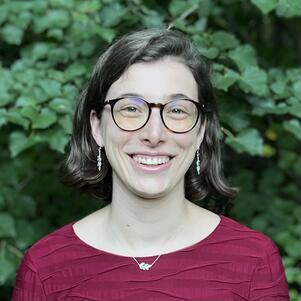 Young woman with chin-length hair and glasses wearing dark red shirt, standing in front of trees