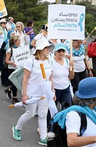 Two women wearing white and marching  holding a sign that says "Women Wage Peace" in English, Hebrew, and Arabic