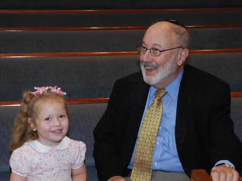 Six year old in pink dress seated next to elderly man dressed in a suit, sitting in pews at a synagogue