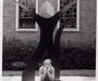 Janet Indick sitting on an outdoor stone plinth, on which sits an abstract statue of curving lines