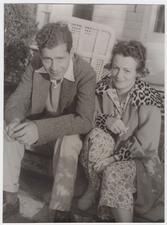 Bella Cohen Spewack and Samuel Spewack sitting on the front porch of a house. Bella Cohen Spewack is holding a cigarette and wearing slippers and a leapord-print housecot over pajamas