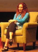 Aline Kominsky speaking on stage, sitting in an armchair