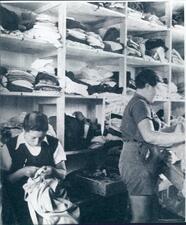 Two women, one sitting and mending a piece and one standing, in the kibbutz clothing warehouse, in front of a wall of shelves with stacks of clothing. 