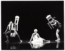 A women in white sits on the ground between two standing people in motion holding chairs in their mouths in Shehechianu: Faith and Science on the Midway