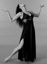 Studio portrait of Susana Weingarten striking a dancing pose, looking up at the ceiling, and wearing a black dress