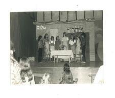 Children in costume standing around a table in front a set decorated like a home with a sign reading shalom in Hebrew behind them for a performance of Fiddler on the Roof