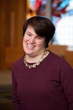 Portrait of Lauren Tuchman smiling in front of a stained glass window wearing a maroon top and gold necklace