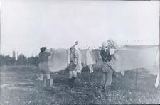 Three women, wearing shorts or pants and boots, hang sheets outside on a line.