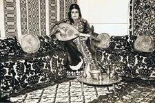 Zohra El Fassia holding an instrument, wearing a dress gifted to her by King Mohammed V, in an ornate room, likely the museum exhibit on North Africa 