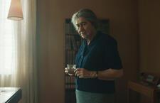 Helen Mirren playing Golda Meir; standing wearing dark shirt, skirt and gray hair pulled back, looking thoughtful