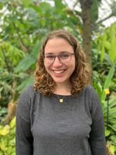 Young woman with brown curly hair and glasses wearing dark gray shirt and posing in front of trees