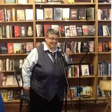 photograph of a woman in a vest speaking at a microphone in a bookstore