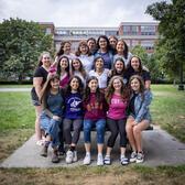 Group photo of the Rising Voices Fellows, sitting on a park bench, park with trees and a building in the background.