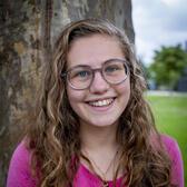 Photo of Rising Voices Fellow Belle Gage, standing in front of a large tree and wearing a pink shirt.