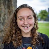 Photo of Isabel Hoffman, in front of a tree wearing a dark blue shirt with an illustration of waves and the sun.