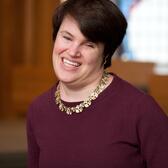 Portrait of Lauren Tuchman smiling in front of a stained glass window wearing a maroon top and gold necklace