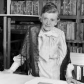 Tatyana Grosman, standing at a table in front of a bookshelf