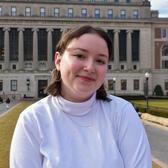 Judy Goldstein has brown hair, brown eyes, and freckles. She is wearing a white turtleneck and standing outside a large building with Ionic columns