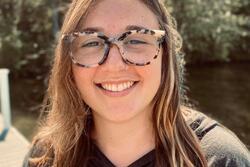 Headshot of Emma Mair with shoulder-length light brown hair and tortoiseshell glasses, posing in front of trees