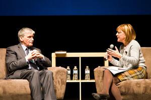 Heather Reisman sitting in an armchair, holding a microphone and speaking to a man seated across from her