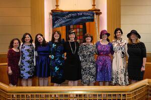 A group of seven women standing at a bimah, posing underneath a banner with Hebrew writing