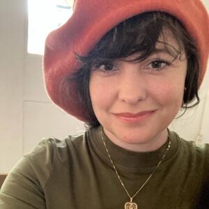 Danielle Kranjec is smiling at the camera, she has short brown hair and brown eyes and is wearing an orange beret and green top with a gold necklace. 