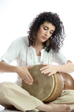 Woman with dark curly hair wearing white blouse and khaki pants sitting and playing drum