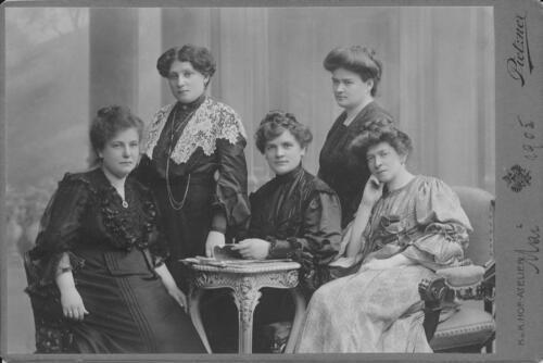 A group of five women in formal dresses, sitting and standing around a small end table