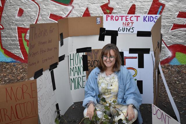 Young woman sitting surrounded by signs: "Protect kids, not guns!" and "Thanks for your thoughts and prayers. How about you fucking do something?"