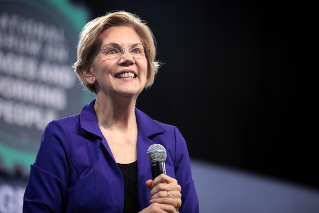 Elizabeth Warren on stage in a purple suit, microphone in hand.