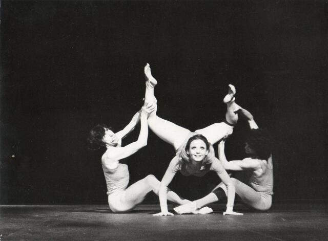 Miguel Angel Palmeros and Bernardo Benitez sitting on a stage, lifting Cecilia Baram's legs in the air as she places her palms on the floor and looks up at the audience