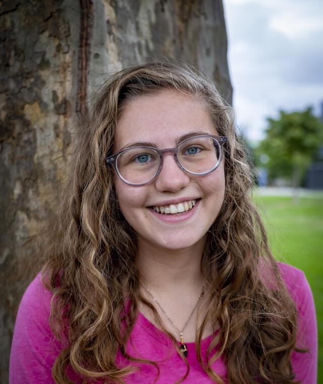 Photo of Rising Voices Fellow Belle Gage, standing in front of a large tree and wearing a pink shirt.