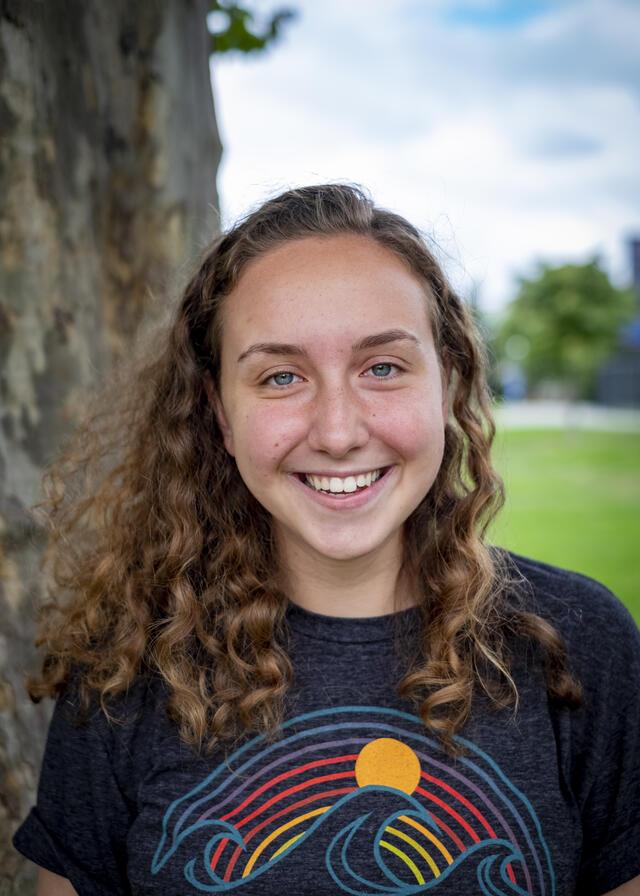 Photo of Isabel Hoffman, in front of a tree wearing a dark blue shirt with an illustration of waves and the sun.