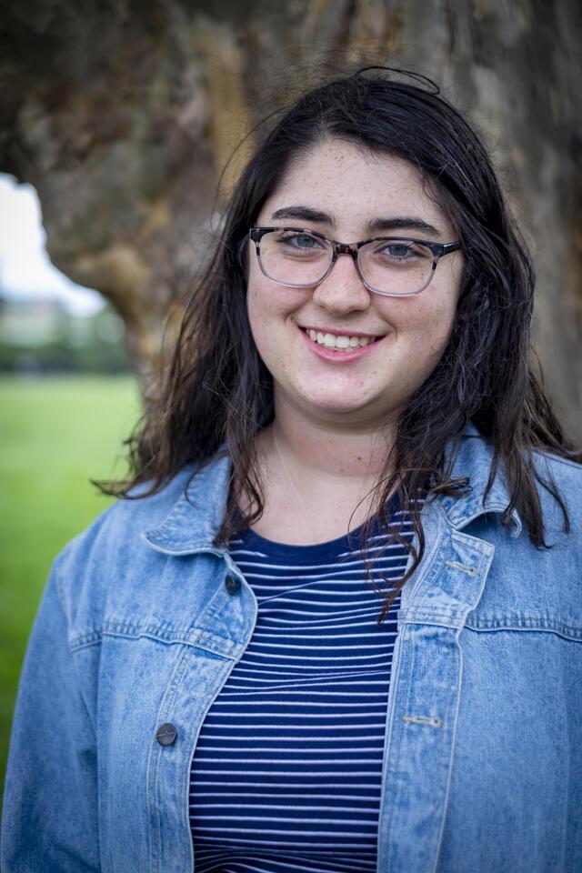 Photo of Lila Goldstein, in front of a tree trunk wearing a striped blue and white shirt and a denim overshirt.