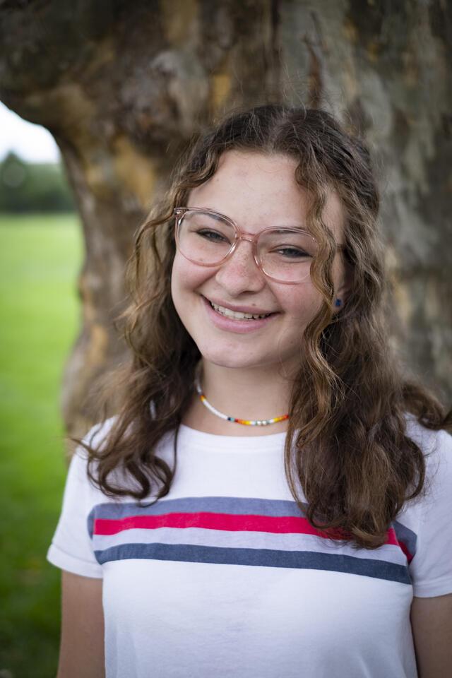 Photo of Neima Fax, in front of a tree wearing a white shirt featuring a multicolor horizontal stripe.