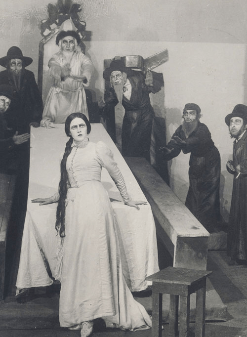 Hanna Rovina onstage, leaning back against a table, surrounded by male actors looking shocked and concerned