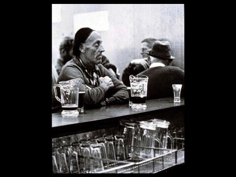 An older man sitting at a bar, with pitchers and pint glasses