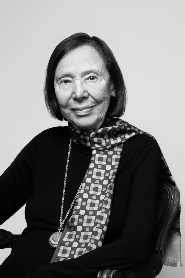 Black and white photo of Grace Schulman, seated in a black outfit with a scarf and locket or pocket watch necklace