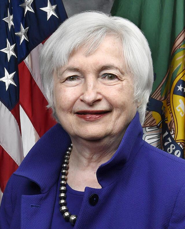 Official portrait of Janet Yellen smiling in a blue jacket and a necklace in front of an American flag