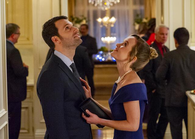 A man and woman in formal attire look up at mistletoe