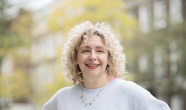 Woman with blond curly hair standing outside, building and trees in background