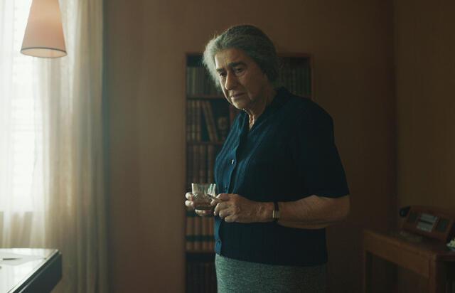 Helen Mirren playing Golda Meir; standing wearing dark shirt, skirt and gray hair pulled back, looking thoughtful