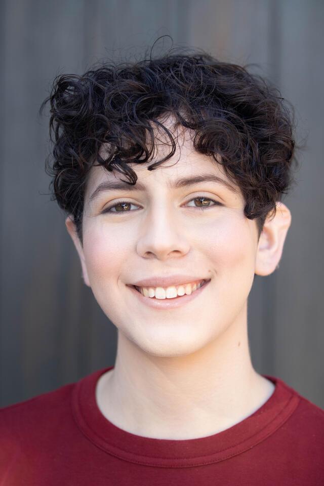 Person with short dark, curly hair and dark red tshirt against dark wall