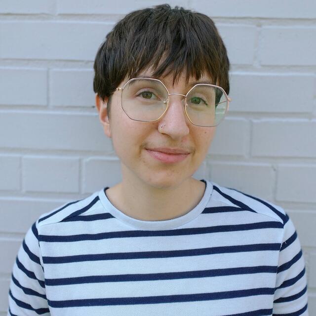 Young person with short brown hair and wire-rimmed glasses wearing a white t shirt with black stripes, posing against a white brick wall